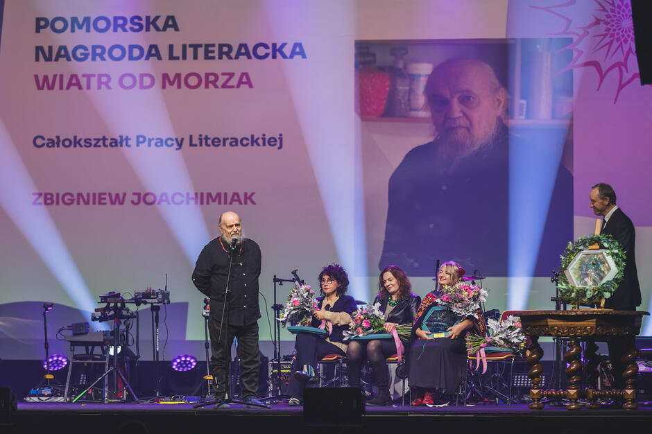 Mężczyzna z długą siwa broda stoi na scenie i przemawia, obok siedza na krzesłąch trzy kobiety z bukietami kwiatów. Obok stoi kolejny mężczyzna bokiem i patrzy na pozostałe osoby. W tle ekran ze zdjęciem mężczyzny przemawiającego i napisem Pomorska Nagroda Literacka Wiatr od morza .