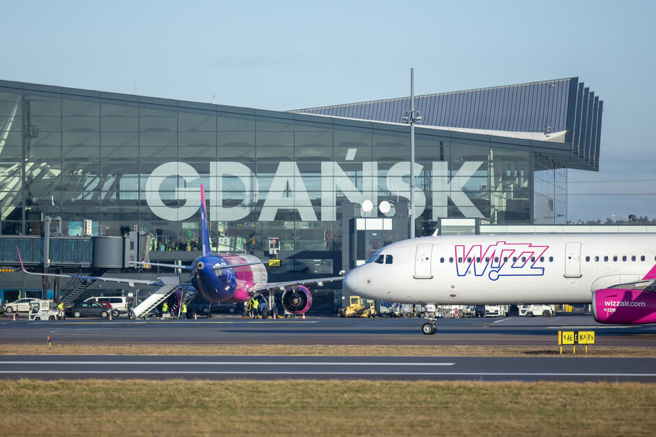Samoloty linii Wizz Air stoją przy terminalu lotniska w Gdańsku, na tle nowoczesnego budynku z dużym napisem „GDAŃSK”. Na płycie lotniska widać obsługę naziemną przygotowującą maszyny do lotu.
