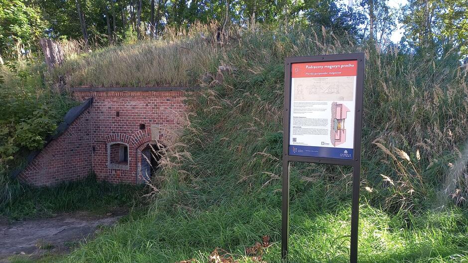 Ceglany budynek częściowo wkopany w ziemię, porośnięty trawą i otoczony zielenią, to dawny podręczny magazyn prochu. Obok znajduje się tablica informacyjna z opisem obiektu i jego przekrojem konstrukcyjnym.
