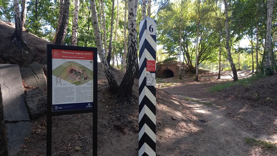 Ścieżka w lesie prowadzi obok tablicy informacyjnej o podręcznym magazynie prochu i biało-czarnego słupa z numerem 6. W tle widać ceglany, sklepiony budynek częściowo wkopany w ziemię, otoczony brzózkami i drzewami liściastymi.
