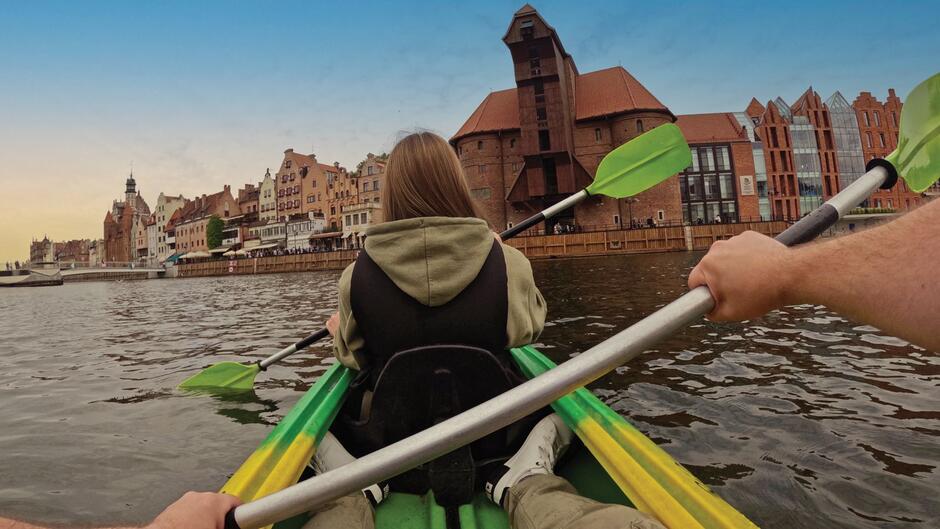 Zdjęcie zrobione z tylnego miejsca w kajaku na rzece. Na pierwszym planie ręce trzymają wiosło, na drugim planie plecy kobiety z wiosłem. W tle stare budynki miasta.