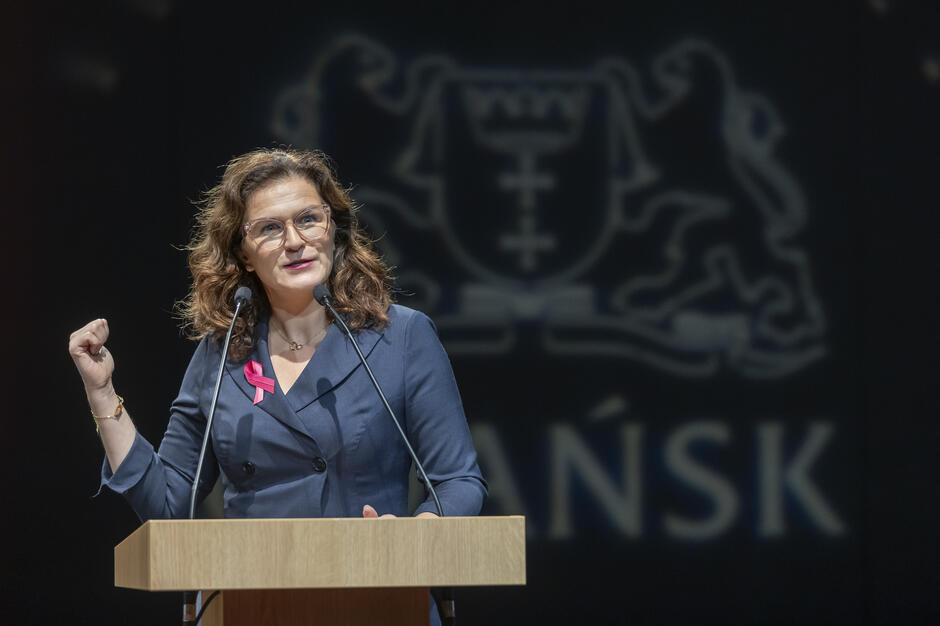 Kobieta przemawia z mównicy, mając za sobą ciemne tło z herbem Gdańska i napisem „GDAŃSK”. Ubrana w granatowy żakiet z różową wstążką, gestykuluje podczas wystąpienia.
