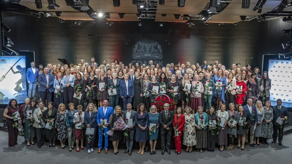 Duża grupa osób pozuje wspólnie do zdjęcia na scenie podczas uroczystości w Gdańsku. Wiele z nich trzyma czerwone róże i dyplomy, co sugeruje wręczenie nagród lub wyróżnień. 