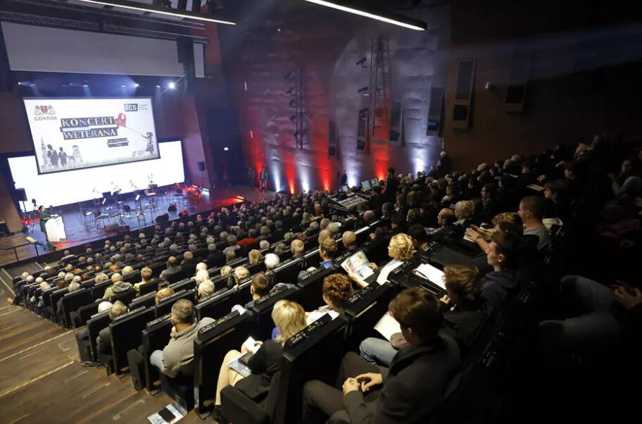 Widok z góry na dużą salę audytoryjną w Europejskim Centrum Solidarności w Gdańsku, wypełnioną publicznością podczas wydarzenia „Koncert Weterana”. Na scenie widać muzyków i osoby przemawiające przy mównicy. W tle duży ekran z napisem „Koncert Weterana” oraz logotypami Miasta Gdańska i ECS. Sala jest oświetlona ciepłym światłem, a ściany podświetlone barwami biało-czerwonymi.