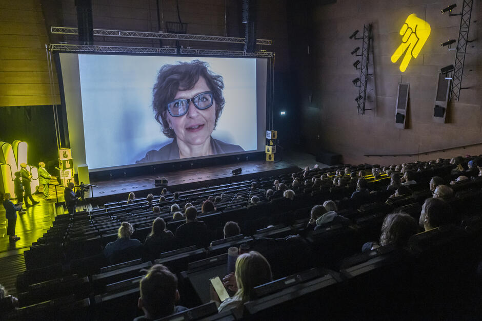 W głębi wielkiej sali - duży telebim z twarzą kobiety w okularach. Przemawia do publiczności która siedzi rządami, w fotelach, w zacienionej części sali 