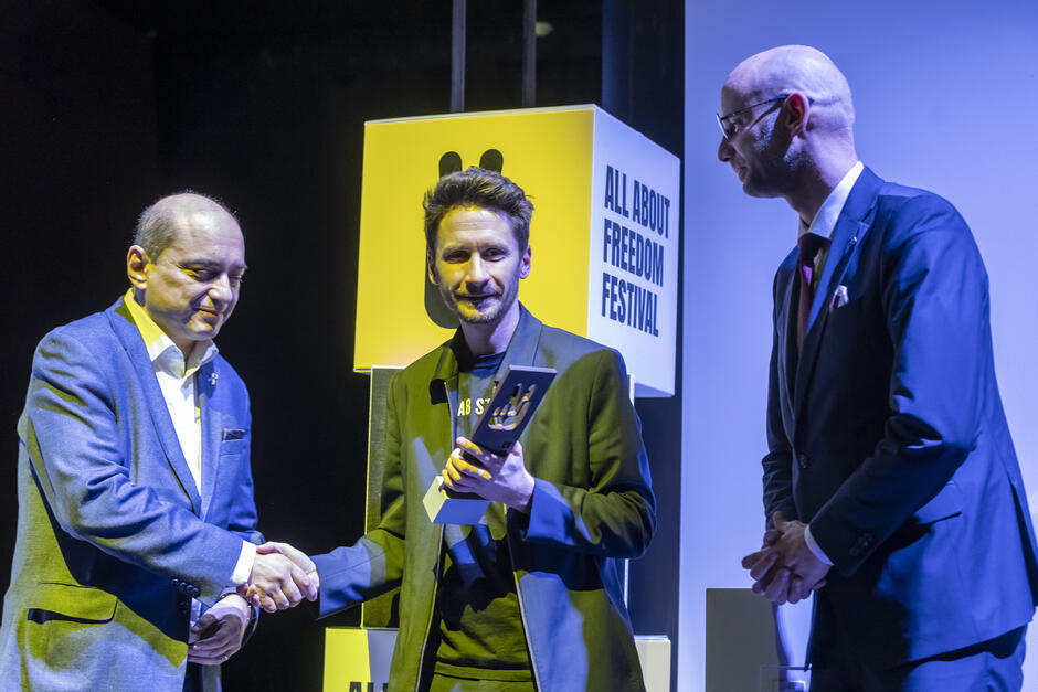 Trzej mężczyźni na scenie, w trakcie przekazywania statuetki