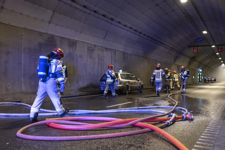 Strażacy wchodzą do tunelu drogowego, prowadząc akcję z wykorzystaniem węży gaśniczych i aparatury tlenowej. Na jezdni widać mokrą nawierzchnię oraz samochód, przy którym prowadzone są działania ratownicze.
