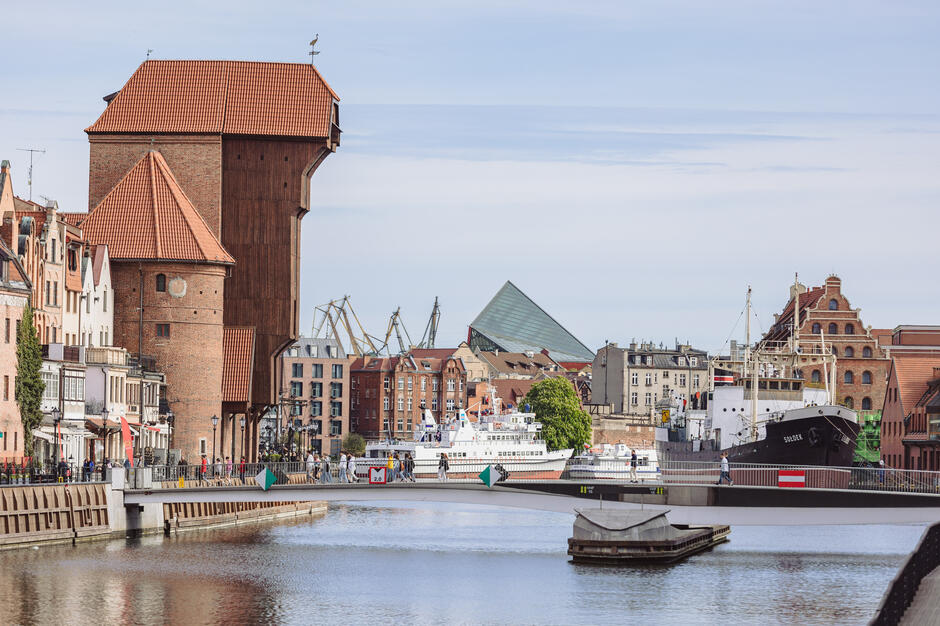 Widok na Długie Pobrzeże w Gdańsku z charakterystycznym Żurawiem nad Motławą, historycznym dźwigiem portowym z XV wieku. W tle widoczne są nowoczesne i zabytkowe budynki oraz statki cumujące przy nabrzeżu, tworzące malowniczy krajobraz miasta. 