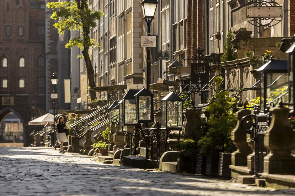 Urokliwa ulica Mariacka w Gdańsku oświetlona porannym słońcem, z charakterystycznymi kamienicami i ozdobnymi schodkami prowadzącymi do domów. Wzdłuż brukowanego chodnika ustawione są stylowe latarnie i rośliny w donicach, tworząc wyjątkowy klimat starego miasta. 