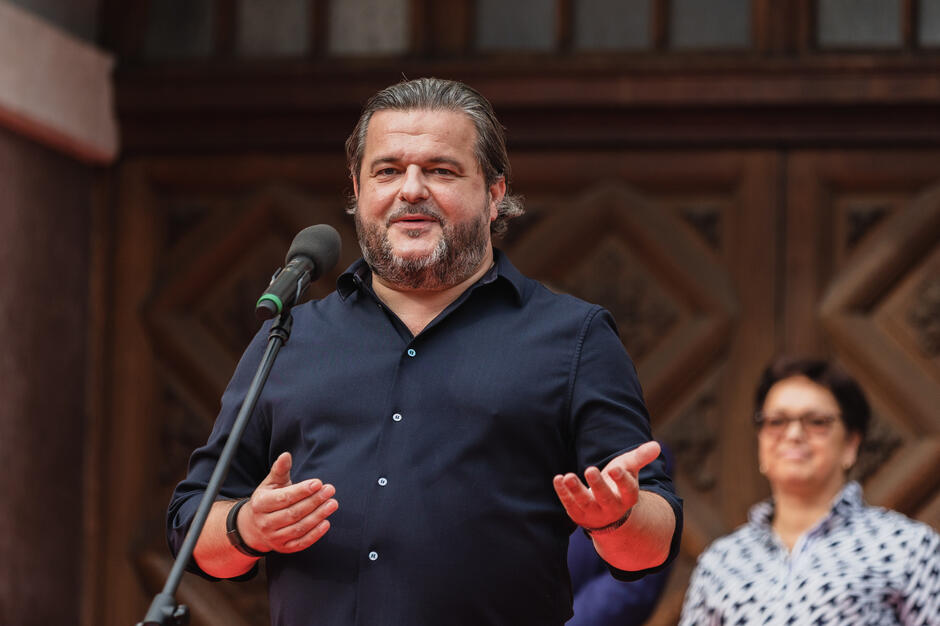 Mężczyzna w ciemnej koszuli przemawia do mikrofonu podczas wydarzenia plenerowego, gestykulując dłońmi. W tle widać ozdobne, drewniane drzwi oraz kobietę obserwującą wystąpienie.
