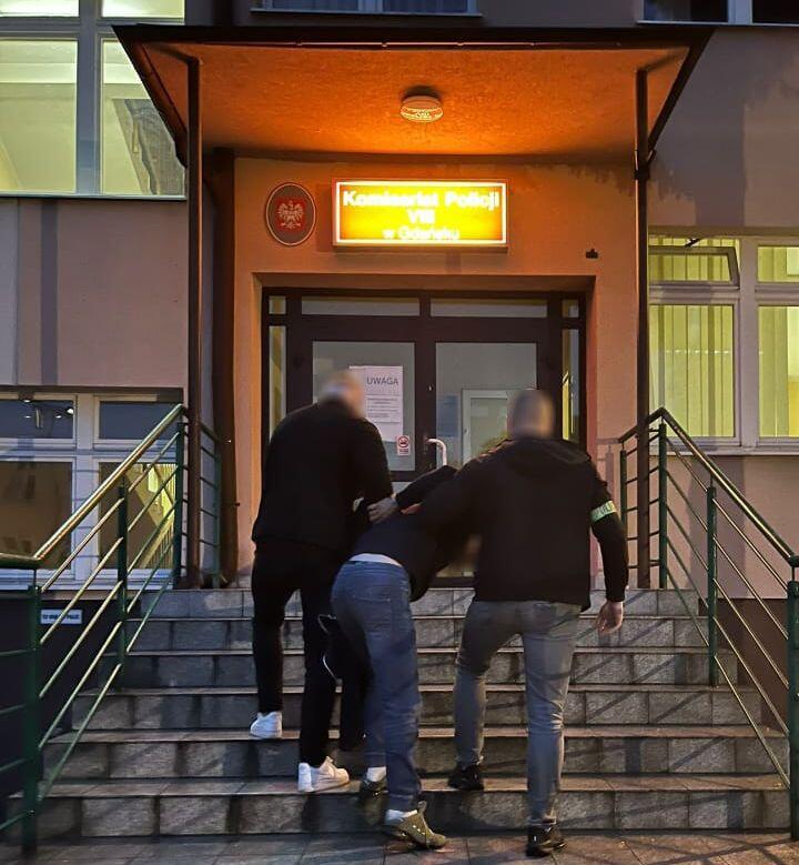Dwóch policjantów w cywilnych ubraniach wprowadza mężczyznę po schodach do komisariatu policji w Gdańsku. Scena odbywa się wieczorem, przed jasno oświetlonym wejściem budynku.