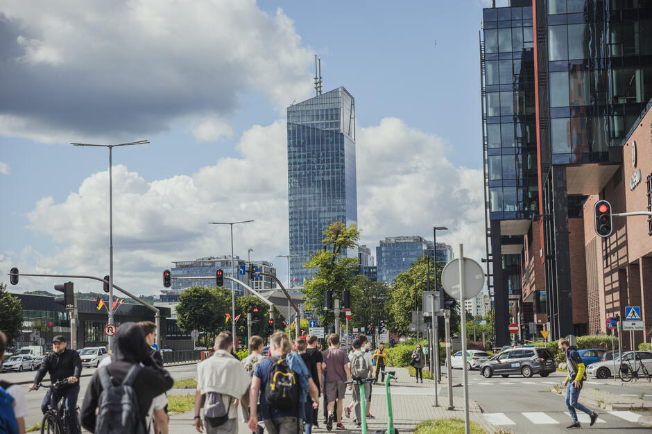 Ruchliwa ulica w Gdańsku z nowoczesnymi biurowcami i pieszymi idącymi w stronę skrzyżowania, nad którym góruje szklany wieżowiec Olivia Star. W kadrze widać także rowerzystów, samochody i sygnalizację świetlną na tle nieba z kłębiastymi chmurami.
