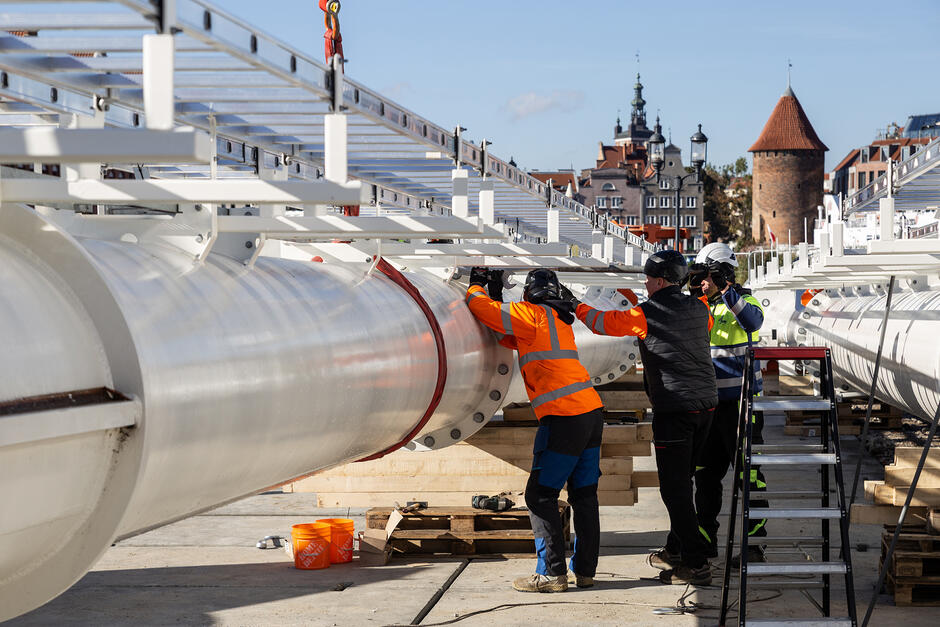 Trzech mężczyzn w kaskach pracuje przy leżącym wielkim stalowym maszcie, w tle widać stare budynki miasta. 