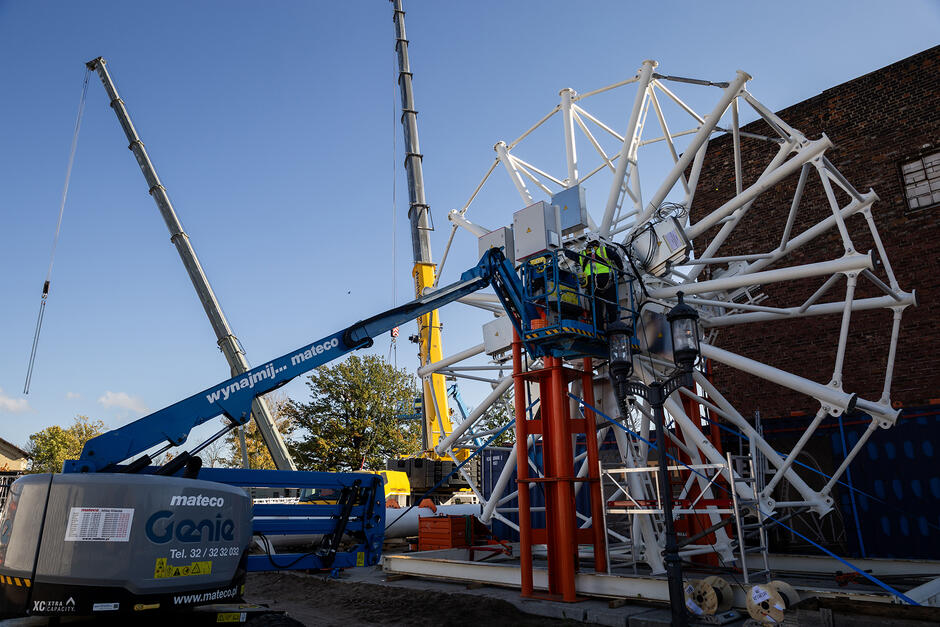 Na placu budowy stoi oś koła widokowego ze szprychami, przy którym pracuje mężczyzna w kasku.