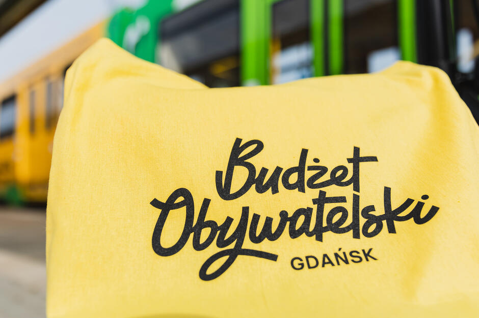 Żółta torba z czarnym tekstem. Tekst brzmi Budżet Obywatelski Gdańsk  , w tle zielono żółty tramwaj