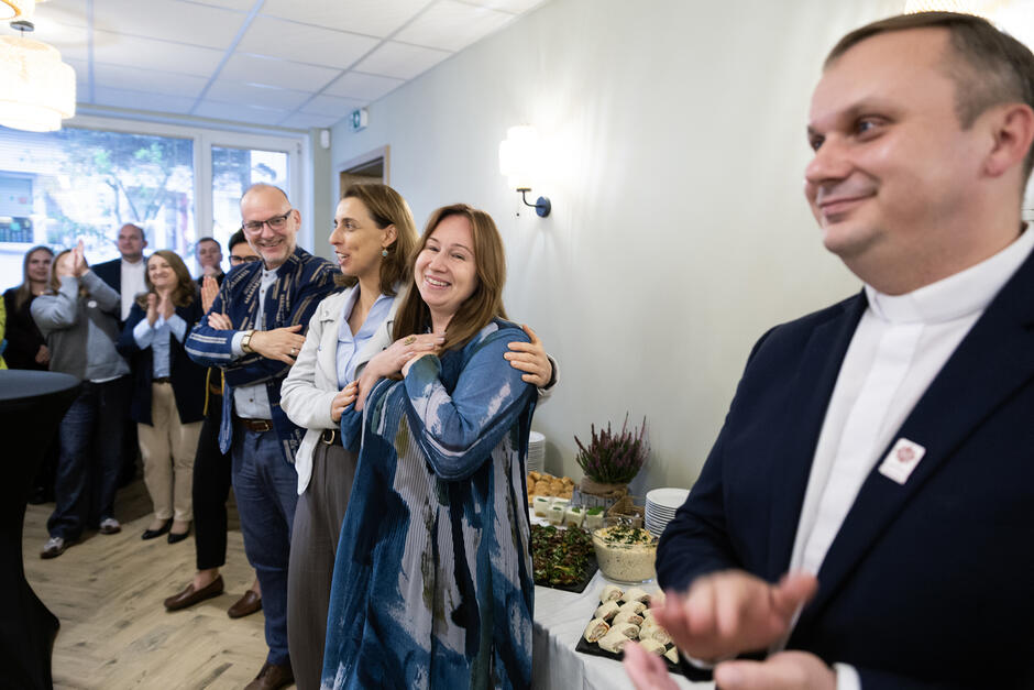 Kilka osób stoi w sali, gdzie odbywa się spotkanie, śmieją się, biją brawo.