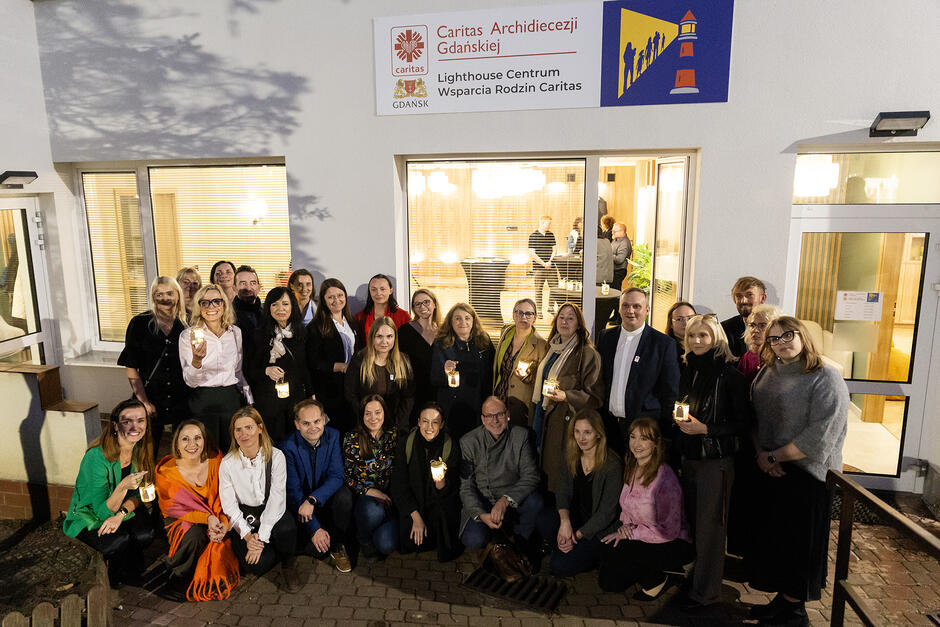 Grupa ludzi pozuje do zdjęcia przed budynkiem. Niektórzy trzymają w rękach lampki.