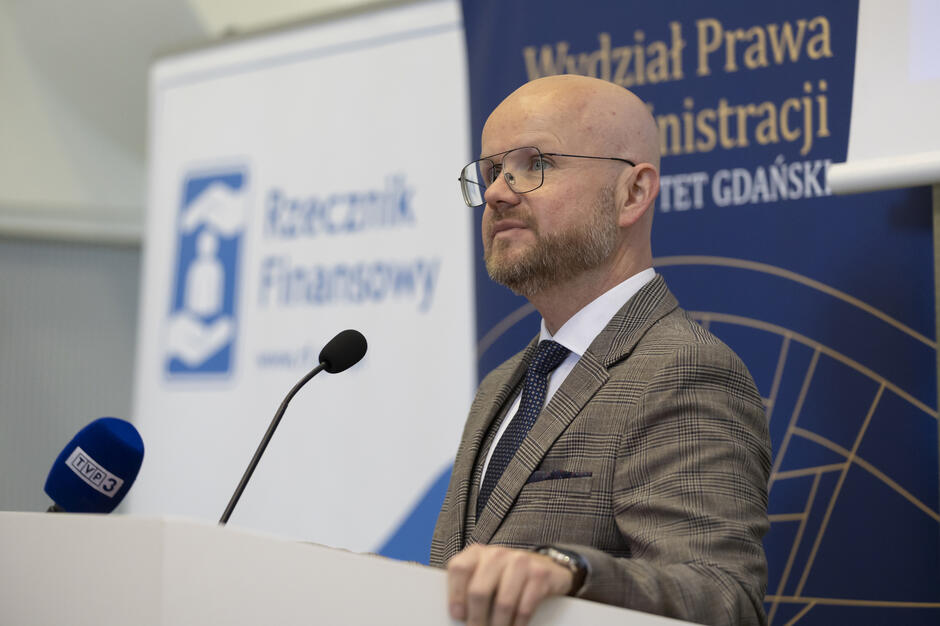 Mężczyzna w garniturze przemawia przy mównicy z mikrofonem podczas oficjalnego wydarzenia zorganizowanego przez Rzecznika Finansowego i Wydział Prawa i Administracji Uniwersytetu Gdańskiego. W tle widoczne są banery instytucji oraz mikrofon stacji TVP3.