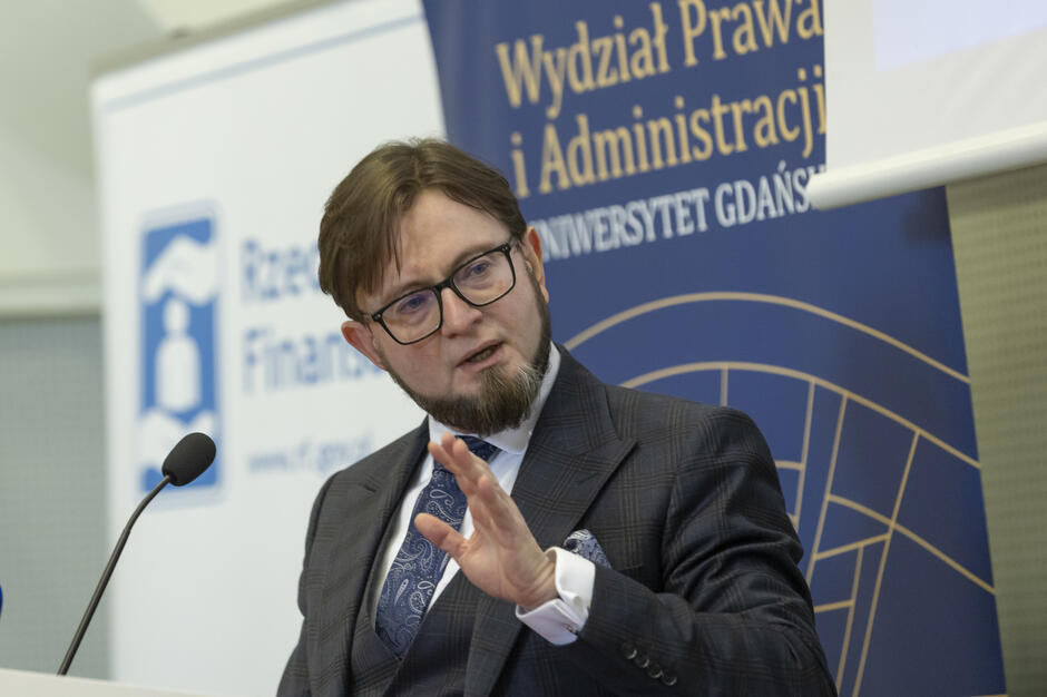 Mężczyzna w garniturze i okularach przemawia podczas konferencji zorganizowanej przez Rzecznika Finansowego oraz Wydział Prawa i Administracji Uniwersytetu Gdańskiego. W tle widoczne są logotypy obu instytucji i mikrofon przy mównicy.