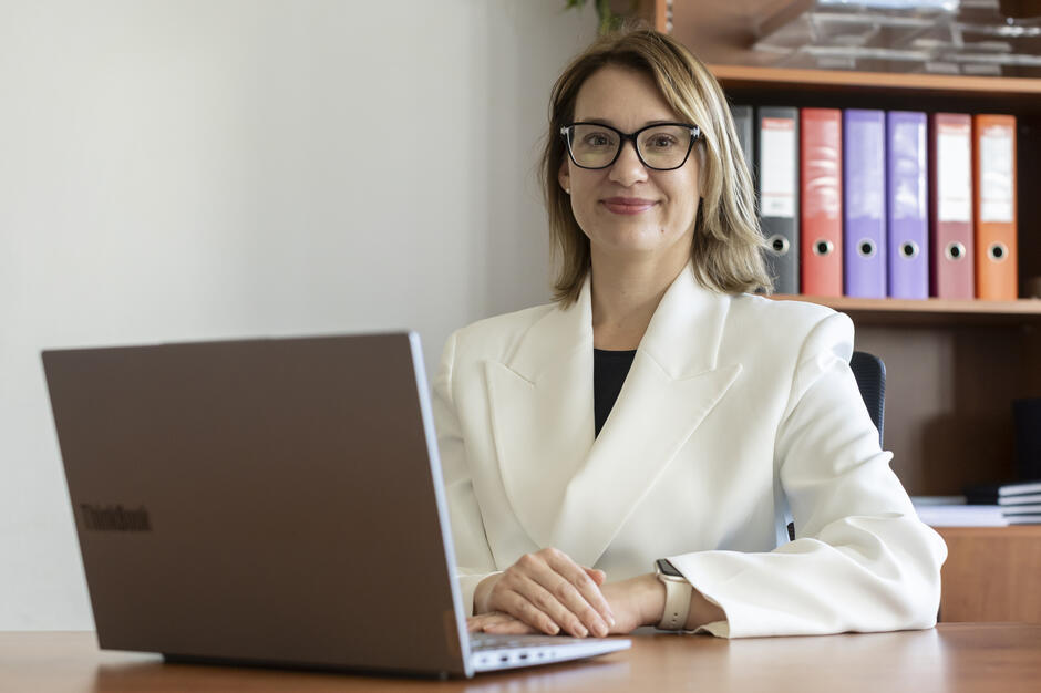 Kobieta w białej marynarce siedzi przy biurku w biurze terenowym Rzecznika Finansowego, przed otwartym laptopem. W tle widać półkę z kolorowymi segregatorami i elementy wyposażenia biura. 