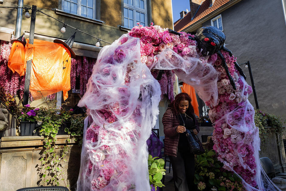 Kobieta wychodzi przez bramkę z lokalu, bramka jest udekorowana halloweenowo, nad kobieta widać wielkiego pająka.