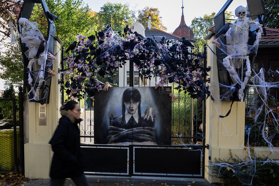 Brama domu z dekoracją halloweenową, obok idzie kobieta.