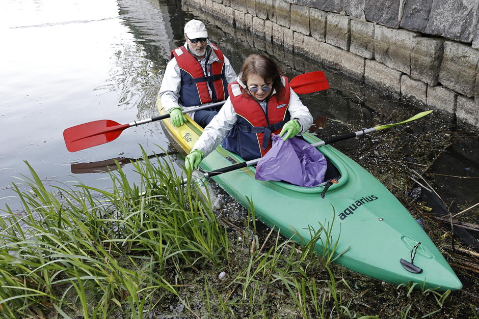 Kobieta i mężczyzna siedzą w kajaku i zbierają przy nabrzeżu szczypcami śmieci z wody do worka plastikowego.