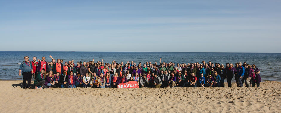 Na plaży nad morzem stoi duża grupa harcerzy i skautów w kolorowych chustach. Wszyscy uśmiechają się do wspólnego zdjęcia, a przed nimi widać czerwony transparent z napisem „Bravely”.