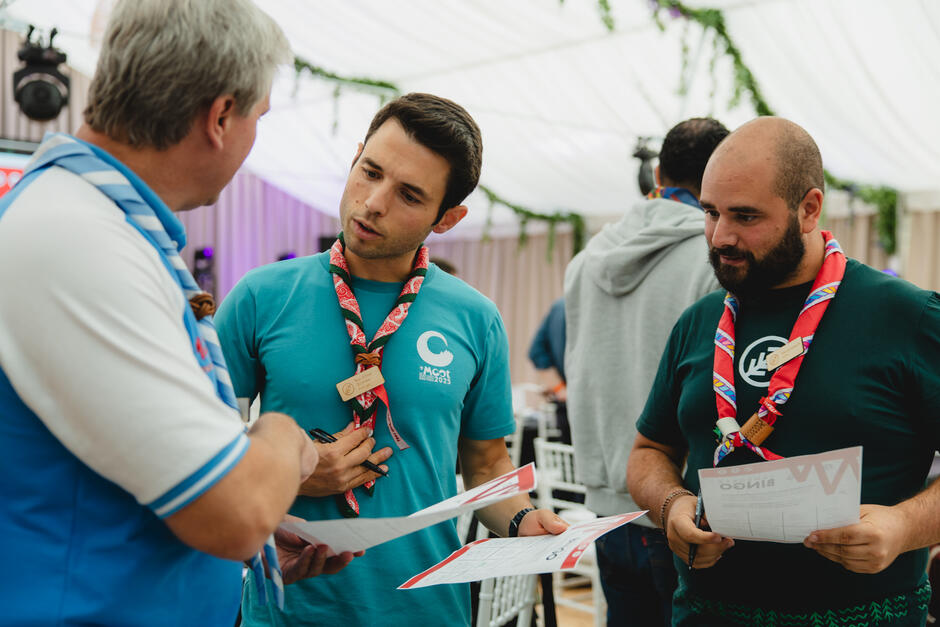 Trzech mężczyzn w harcerskich chustach rozmawia ze sobą w namiocie konferencyjnym. Dwóch z nich trzyma w rękach dokumenty i omawia zapiski.