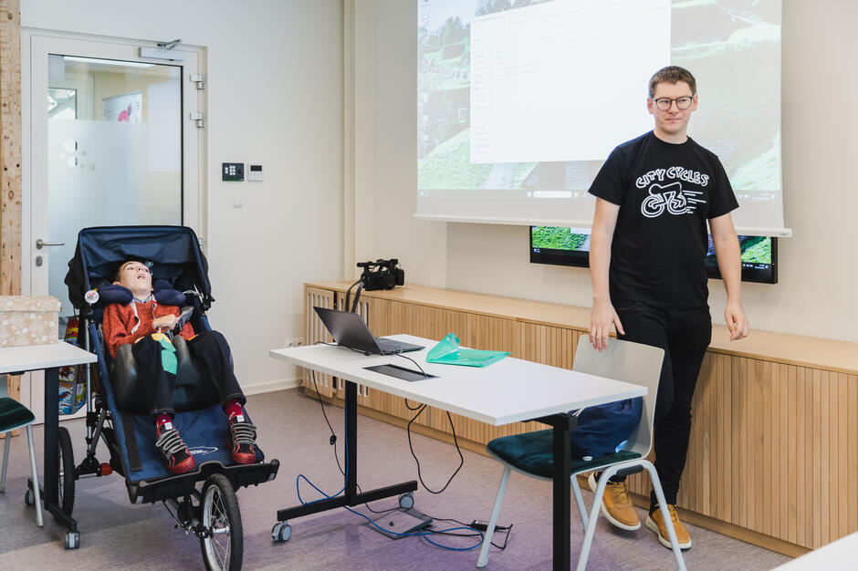 Na zdjęciu widać mężczyznę stojącego obok stołu z laptopem, który prowadzi prezentację w jasnym pomieszczeniu. Obok niego znajduje się chłopiec w wózku inwalidzkim, uważnie uczestniczący w spotkaniu.
