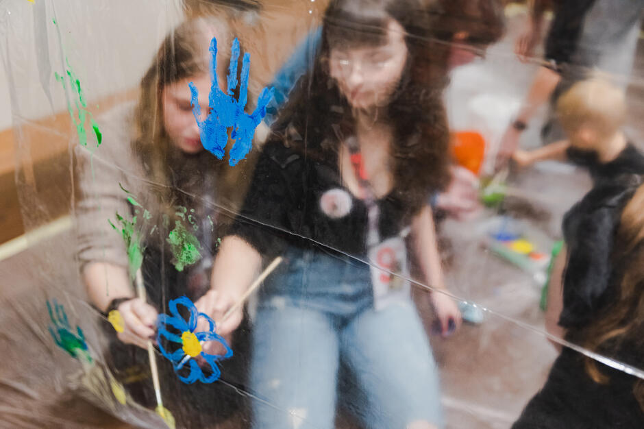 Na zdjęciu widać osoby malujące kolorowe wzory farbami na przezroczystej folii, tworzące kwiaty i odciski dłoni. W tle uczestnicy warsztatów skupieni są na wspólnej, kreatywnej pracy plastycznej.
