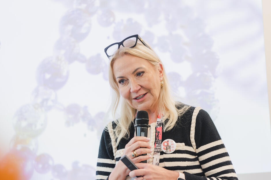 Na zdjęciu kobieta z mikrofonem w dłoni przemawia podczas wydarzenia, mając na szyi identyfikator i przypinkę. W tle widać rozmyty obraz przedstawiający kuliste formy, prawdopodobnie element prezentacji multimedialnej.
