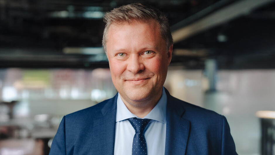 Na zdjęciu mężczyzna w garniturze i krawacie patrzy w obiektyw z lekkim uśmiechem. Tło jest rozmyte, co podkreśla formalny i profesjonalny charakter portretu. 