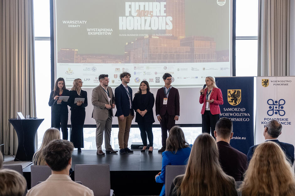 Kilka osób stoi na scenie w czasie konferencji, przed nimi siedzą ludzie na krzesłach, za nimi ekran z napisem Future Horizons 2025 .