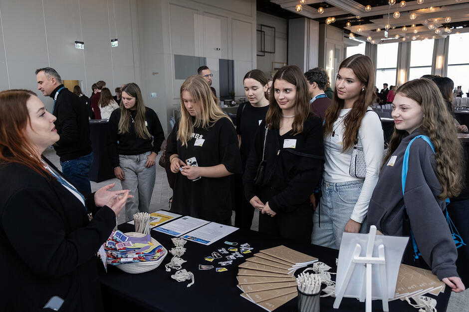 Młode kobiety, uczennice stoją przy stanowisku firmy podczas konferencji i rozmawiają z kobietą na stoisku.