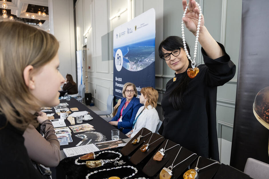 Kobieta na stoisku na konferencji pokazuje swoje wyroby rzemieślnicze - biżuterię z bursztynem.