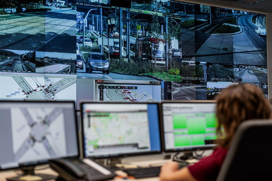 Pracownik centrum zarządzania ruchem monitoruje na ekranach sytuację na ulicach miasta, korzystając z systemu kamer i map drogowych. Na dużym ekranie widać podgląd z wielu kamer obejmujących różne skrzyżowania i trasy w Gdańsku.
