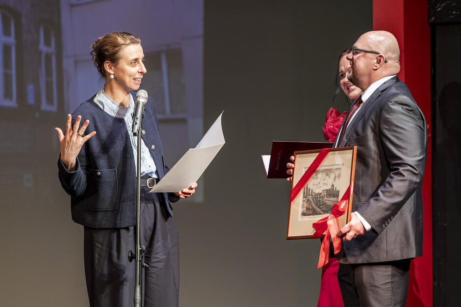 Podczas uroczystości kobieta przemawia przy mikrofonie, trzymając kartkę z notatkami. Obok niej mężczyzna w garniturze odbiera oprawiony w ramkę obraz przewiązany czerwoną wstążką.
