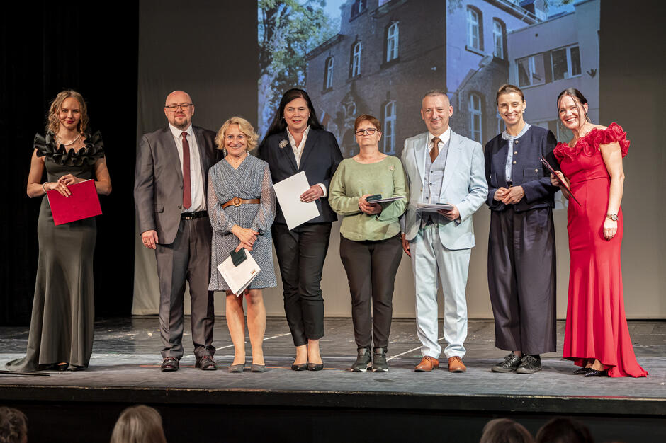 Grupa osób pozuje na scenie podczas uroczystego wydarzenia, trzymając w dłoniach dyplomy i dokumenty. Uczestnicy są elegancko ubrani, a w tle widać projekcję przedstawiającą budynek związany z okazją spotkania.
