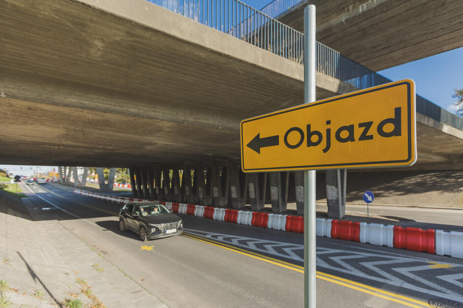 Znak drogowy informuje o objeździe w lewo, ustawiony przy jezdni pod betonową estakadą. Na drodze widać samochód osobowy oraz barierki oddzielające pas ruchu w trakcie prac drogowych. 