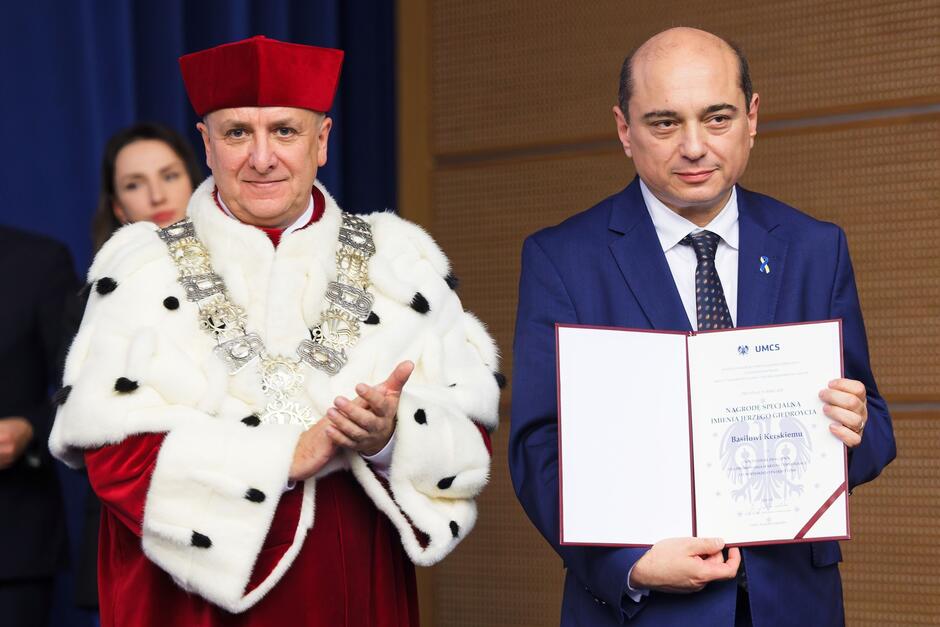 Dwaj mężczyźni na scenie: ten po prawej jest ubrany w garnitur, trzyma w rękach dyplom; ten po prawej ubrany jest w strój rektora uczelni, z gronostajami