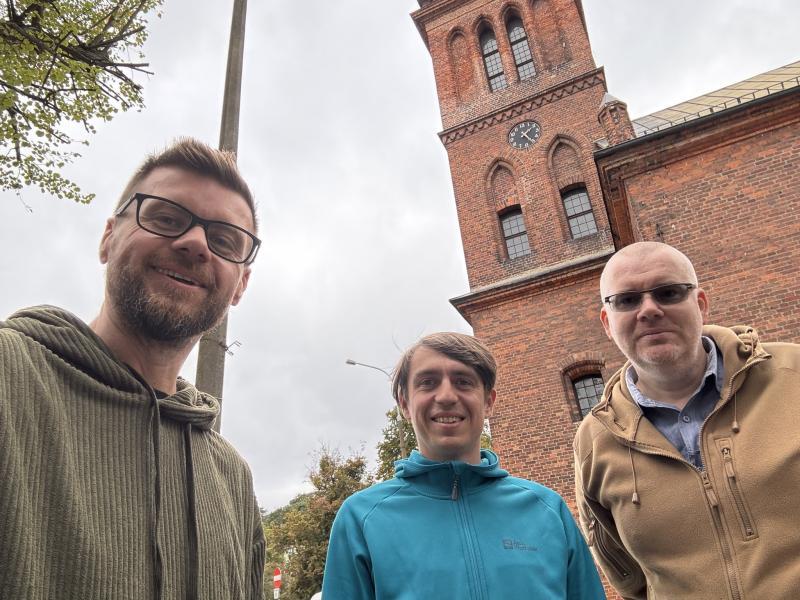 Trzech mężczyzn stoi przed ceglanym budynkiem z wieżą zegarową, patrząc w stronę aparatu i uśmiechając się. Scena odbywa się w pochmurny dzień, a w tle widać drzewa i fragment ulicznego oświetlenia. 