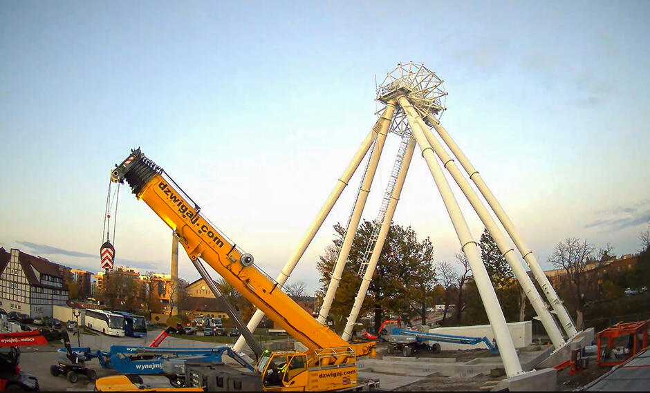 Na placu budowy stoi sześć masztów, na których zamontowana jest oś koła widokowego. Obok stoi dźwig.