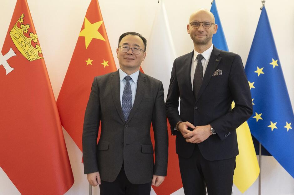 Chińczyk i Polak na tle stojących flag niemal 2-metrowej wysokości. Chińczyk jest po lewej