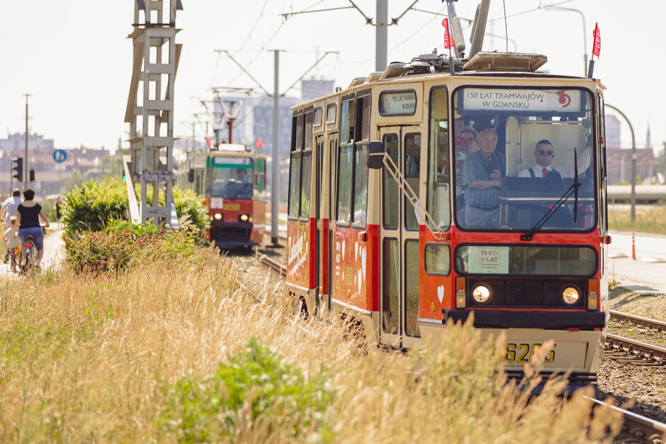 Zabytkowy tramwaj z napisem „150 lat tramwajów w Gdańsku” jedzie po torach w słoneczny dzień, a w środku widać pasażerów i motorniczego w eleganckim stroju. W tle zbliża się kolejny pojazd szynowy, a obok torowiska poruszają się rowerzyści.
