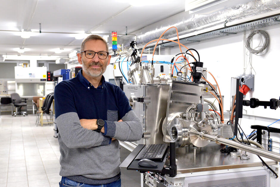 Mężczyzna w okularach stoi z założonymi rękami w nowoczesnym laboratorium badawczym, obok zaawansowanego urządzenia z licznymi przewodami i metalowymi elementami. W tle widoczne są stanowiska pracy, sprzęt laboratoryjny i jasne oświetlenie pomieszczenia. 