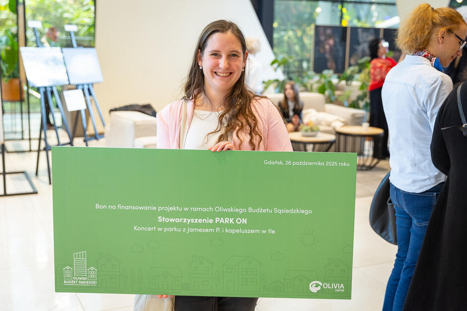 Kobieta w jasnym stroju trzyma duży zielony bon symbolizujący finansowanie projektu w ramach Oliwskiego Budżetu Sąsiedzkiego dla Stowarzyszenia PARK ON. W tle widać uczestników wydarzenia rozmawiających w nowoczesnym, jasno oświetlonym wnętrzu. 