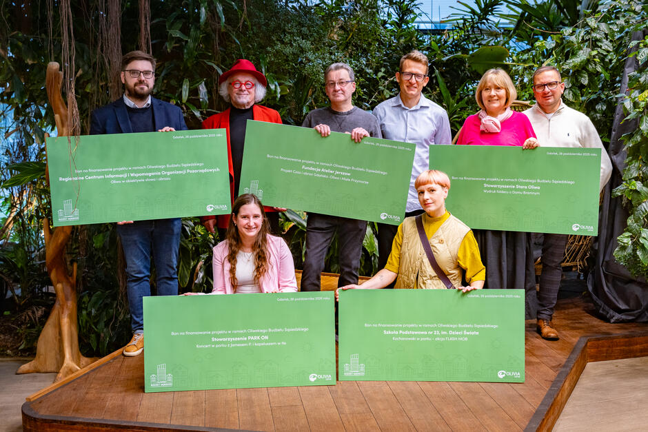Grupa osób pozuje z dużymi zielonymi tablicami symbolizującymi bony na finansowanie projektów w ramach Oliwskiego Budżetu Sąsiedzkiego. Zdjęcie wykonano w otoczeniu roślin, a uczestnicy uśmiechają się, prezentując różnorodne inicjatywy lokalne. 