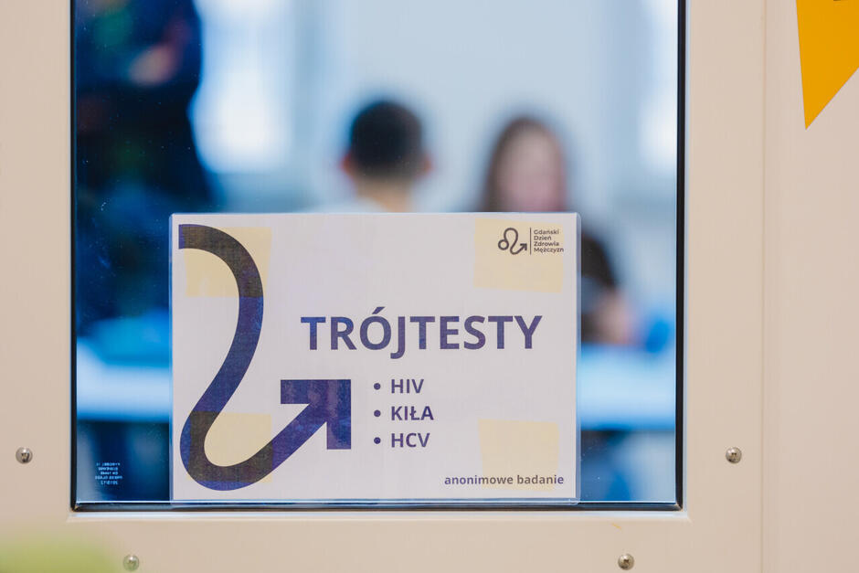 Zdjęcie przedstawia przyklejoną do szyby kartkę z napisem ‘TRÓJTESTY HIV, KIŁA, HCV – anonimowe badanie’. W prawym górnym rogu widnieje logo Gdańskiego Dnia Zdrowia Mężczyzn. Za szybą, w nieostrym tle, widać jedną osobę stojącą oraz dwie siedzące przy stole w jasnym pomieszczeniu.”