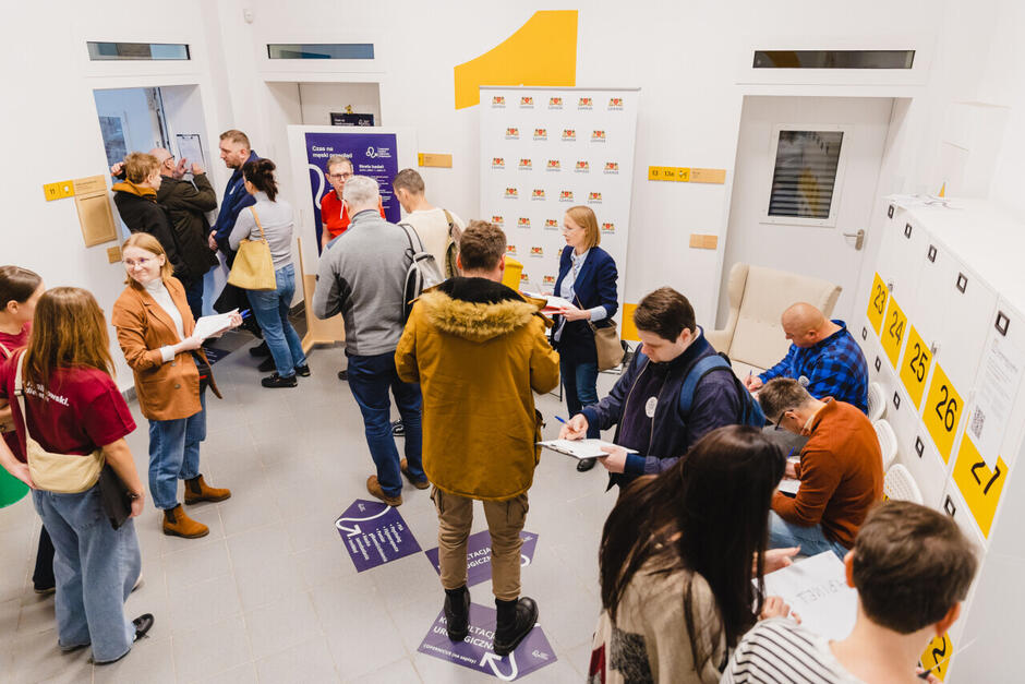 Uczestnicy pierwszej edycji Gdańskiego Dnia Zdrowia Mężczyzn wypełniają ankiety i rozmawiają w jasnym korytarzu Centrum Dolna Brama. W tle widoczne są plakaty informacyjne, szafki z numerami oraz oznaczenia na podłodze.